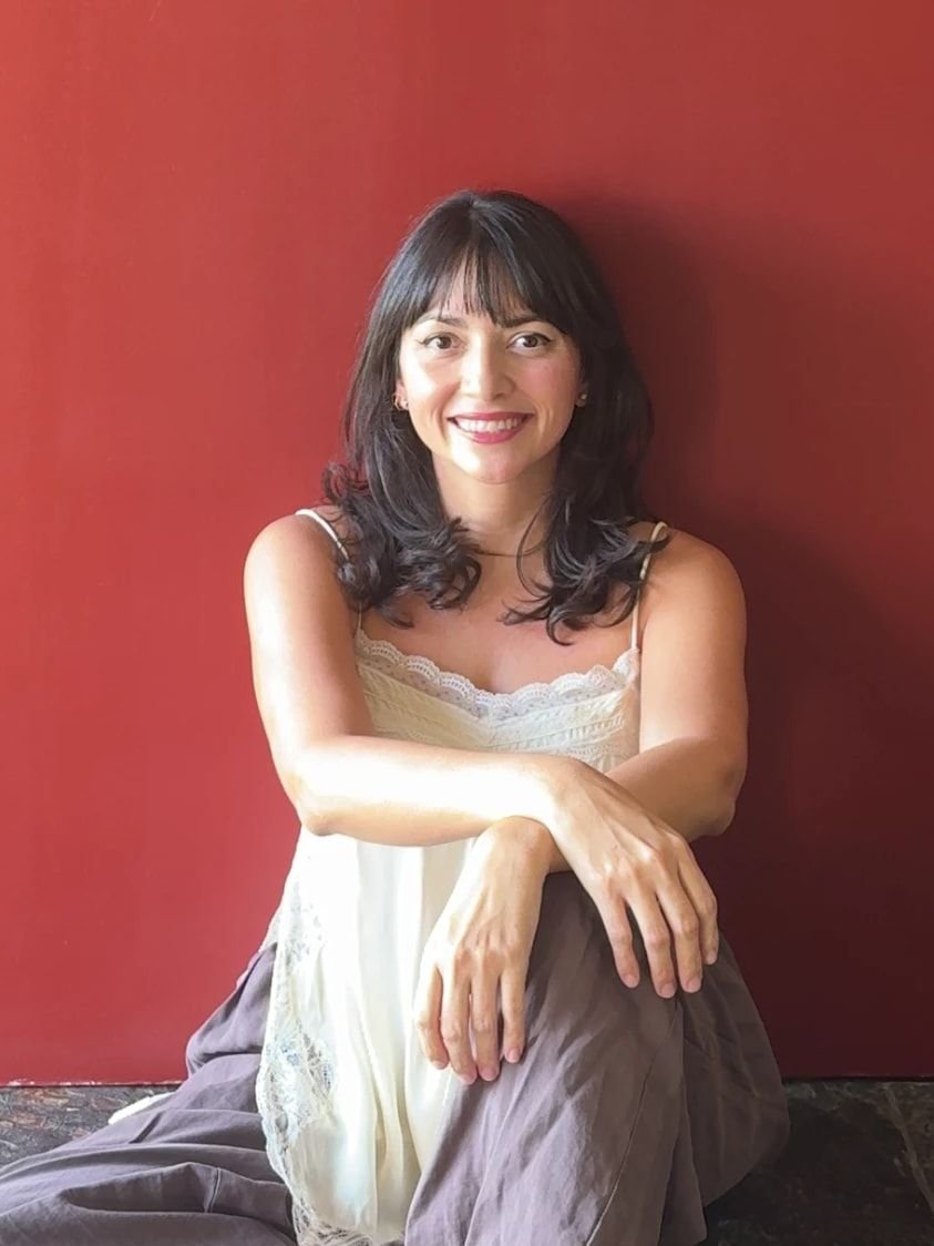 Alexandra Arce sonriendo, sentada frente a una pared roja con los brazos apoyados en la rodilla