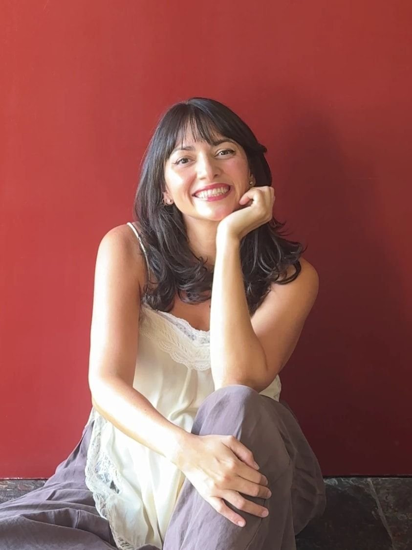 Alexandra Arce sonriendo con la mano en la barbilla, frente a una pared roja